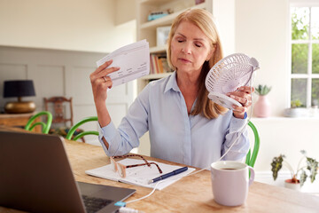 Menopausal Mature Woman Having Hot Flush At Home Cooling Herself With Fan Connected To Laptop