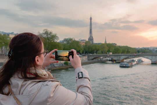 Young woman taking picture with the cellphone from the Eifel tower at sunset. With bridge and the Seine river in Paris.