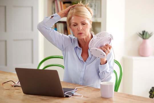 Menopausal Mature Woman Having Hot Flush At Home Cooling Herself With Fan Connected To Laptop