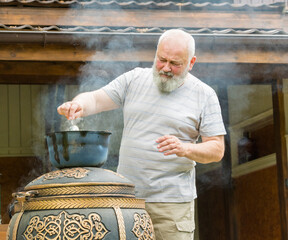 an elderly man cooks pilaf in a pot on a tandoor stove.