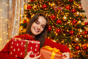 Pleased smiling girl sitting near decorated Christmas tree with present boxes in her hands. Magical happy time of winter holidays and warm atmosphere