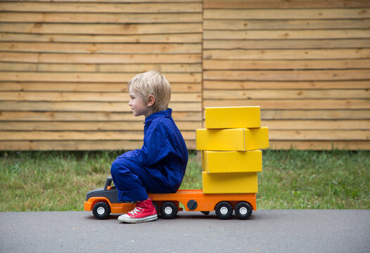 6-year-old boy in a blue overalls - uniform sits on a large toy car - a truck with a lot of yellow cardboard boxes. Parcel delivery, postman, little truck driver. Positive, humor, joy