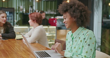 A happy black woman using laptop at coffee shop. One African American girl typing on computer keyboard smiling. A female entrepreneur person looking at laptop screen