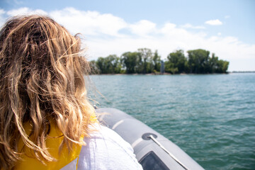 A young woman with windblown hair looks out at a forested island on the Canadian side of Lake Erie near Pelee Island from a Zodiac boat during the summertime. Clear, calm water, blue skies, sunshine.