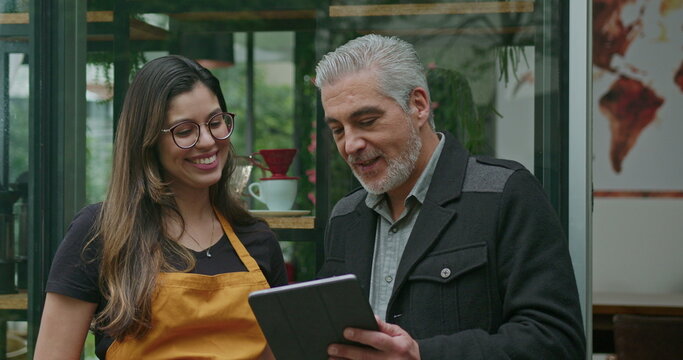 Small business owner speaking with female employee barista wearing apron. Boss manager talking with staff explaining restaurant business holding tablet device