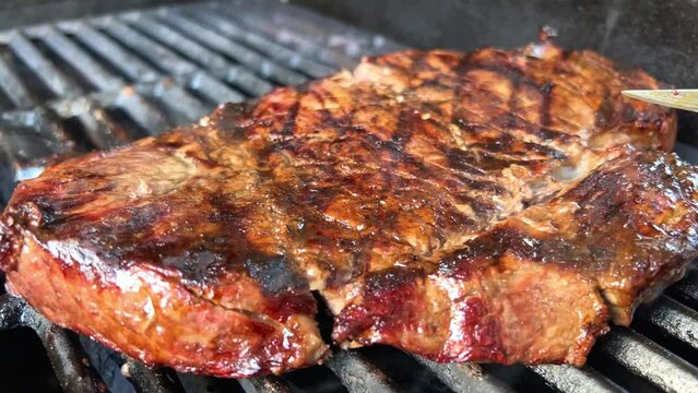 a huge piece of meat on a barbecue grill is lifted with a fork and turned over it is already cut in several places with a knife it is very appetizing medium roasting inside there is a lot of blood