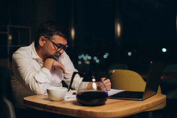 Caucasian boss working late sitting on desk in office at night. Business man feeling tired and stress for overload job..