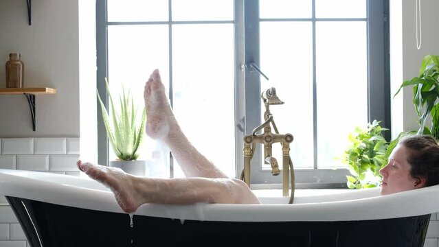 woman in a bathroom with a large window puts her feet on the edge of the tub