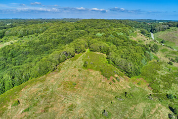 Springtime. Aerial view of a field at the top of a hill in the countryside, surrounded by green forestry and trees on a sunny spring or summer day. Scenery of nature with the blue sky on the horizon.