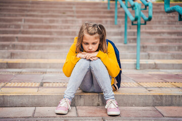 Back to elementary, primary school. Little sad unhappy girl with backpack. Lonely schoolgirl with emotional problems, victim of bullying in schoolyard. Teen in depresiion sitting alone on stair steps