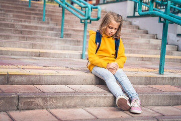 Back to elementary, primary school. Little sad unhappy girl with backpack. Lonely schoolgirl with emotional problems, victim of bullying in schoolyard. Teen in depresiion sitting alone on stair steps