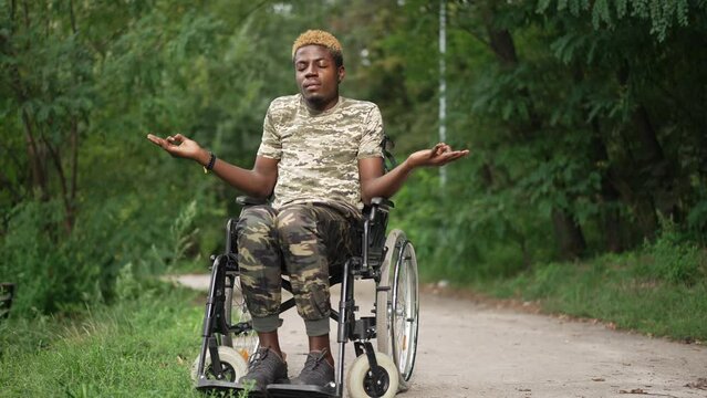 Brave African American man in wheelchair meditating sitting in summer park outdoors with closed eyes. Wide shot portrait of calm courageous young soldier calming down outdoors thinking. Slow motion