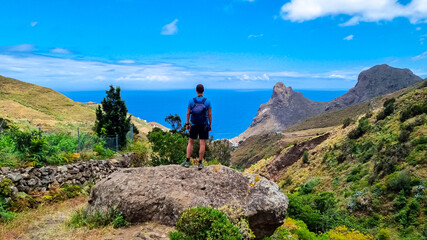 Backpack man with scenic view of Atlantic Ocean coastline and Anaga mountain range on Tenerife, Canary Islands, Spain, Europe. Looking at Roque de las Animas crag. Hiking trail from Afur to Taganana