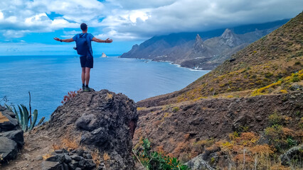 Fototapeta premium Backpack man with scenic view of Atlantic Ocean coastline and Anaga mountain range on Tenerife, Canary Islands, Spain, Europe. Looking at Roque de las Animas crag. Hiking trail from Afur to Taganana