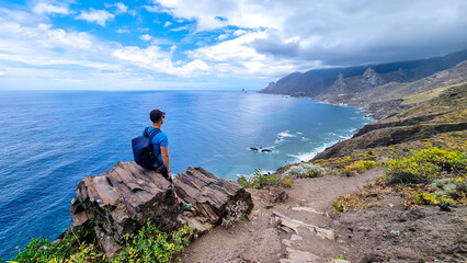 Backpack man sitting on rock with scenic view of Atlantic Ocean, Anaga mountains, Tenerife, Canary Islands, Spain, Europe. Looking at Roque de las Animas crag. Hiking trail from Afur to Taganana