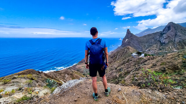 Backpack man with scenic view of Atlantic Ocean coastline and Anaga mountain range on Tenerife, Canary Islands, Spain, Europe. Looking at Roque de las Animas crag. Hiking trail from Afur to Taganana