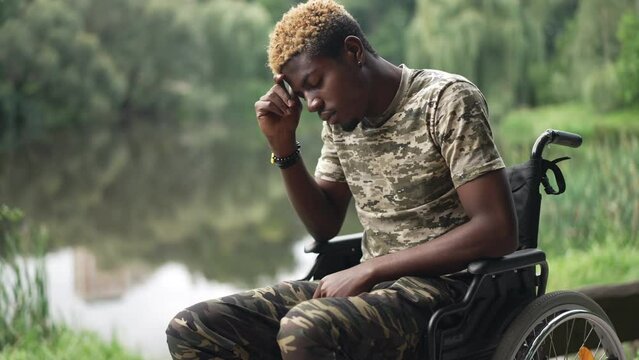 Side view devastated African American man in wheelchair thinking sitting in park on lake shore. Sad depressed frustrated soldier resting outdoors on summer day. Procrastination and disability concept