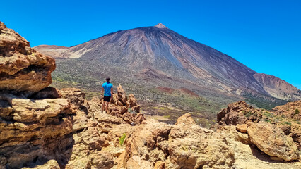 Fototapeta na wymiar Sport man on summit of Riscos de la Fortaleza with panoramic view on volcano Pico del Teide, Mount El Teide National Park, Tenerife, Canary Islands, Spain, Europe. Close up view on rock in foreground