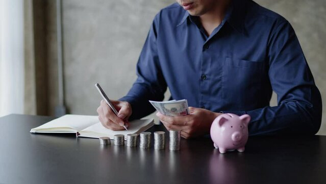Close up of business man’s hands holding money and writing on the notes on the table with stack of coins and piggy savings.