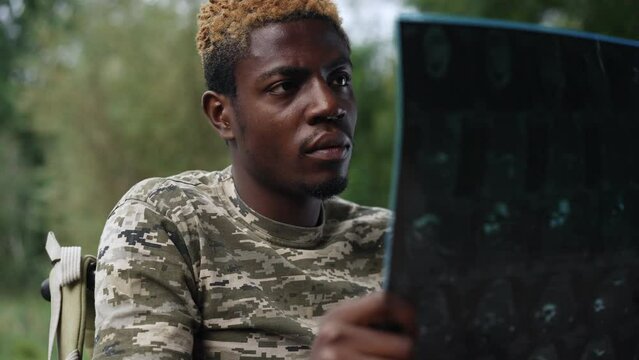 Portrait of depressed sad man in wheelchair with X-ray thinking outdoors. African American young handsome frustrated guy analyzing medical result resting in park forest. Slow motion