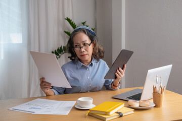 Portrait of an elderly woman working inside the house using laptops and tablets to work and check important documents for customers.