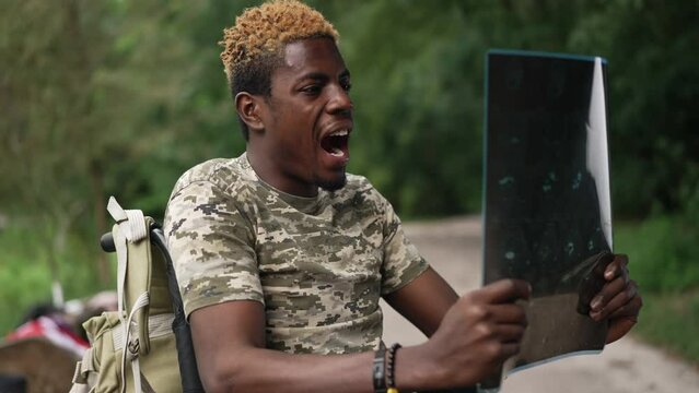 Devastated African American soldier in wheelchair screaming examining X-ray outdoors. Side view portrait of hopeless young ill man freaking out in park. Nervous breakdown and war consequences PTSD