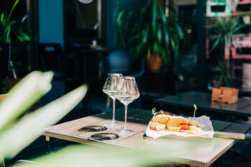A wooden table in a restaurant with a cheese plate, grape, honey bread and white wine. Wine glasses and cheese served for a party in a blurred bar or a restaurant on terrace at a sunny summer day.