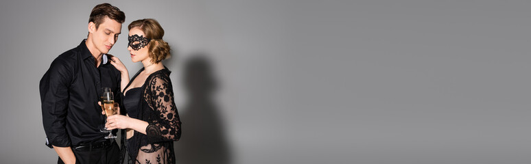 hot woman in lace mask and tunic clinking champagne glasses with man on grey background, banner.
