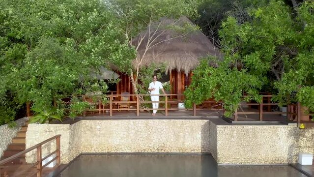Aerial shot flying away from a young man drinking and celebrating life in a bungalow house with oceanfront view