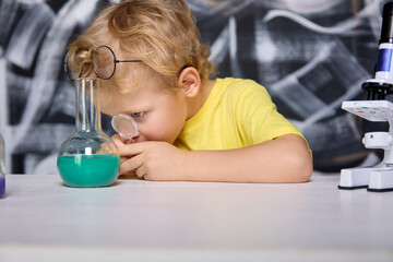 Blonde boy in glasses carefully examines through magnifying glass green liquid in glass tube. Chemical substance in flask aroused genuine interest in the young scientist in the children's laboratory