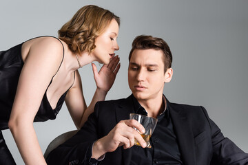 sexy woman whispering in ear of man with glass of whiskey isolated on grey.