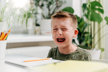 Back to school. Crying school kid boy studying at home and doing homework. Sad caucasian child siting at table with notepad, pencils and training books. Distance learning online education.