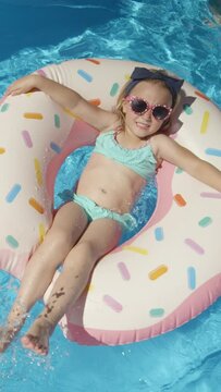 TOP DOWN: Cute little girl with sunglasses on a floatie donut in swimming pool. Cheerful girl having fun floating and making water splashes with her legs. Happy kid enjoying summer in backyard pool.
