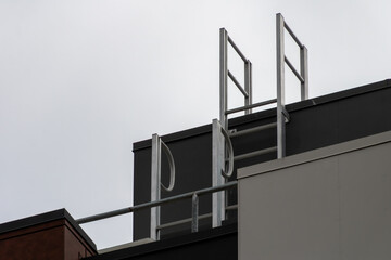 A grey and black building showing a metal built in ladder going to the roof. A metal rail can be seen on the left. A grey sky is in the background.
