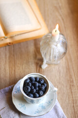 Vintage porcelain cup filled with fresh blueberries, pressed flowers, open book, reading glasses and lit candle on the table. Hygge at home. Selective focus.