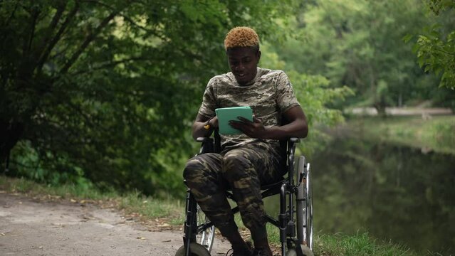 Smiling African American man in wheelchair surfing social media on tablet sitting on lake shore in summer park. Wide shot portrait of positive confident relaxed soldier enjoying leisure outdoors