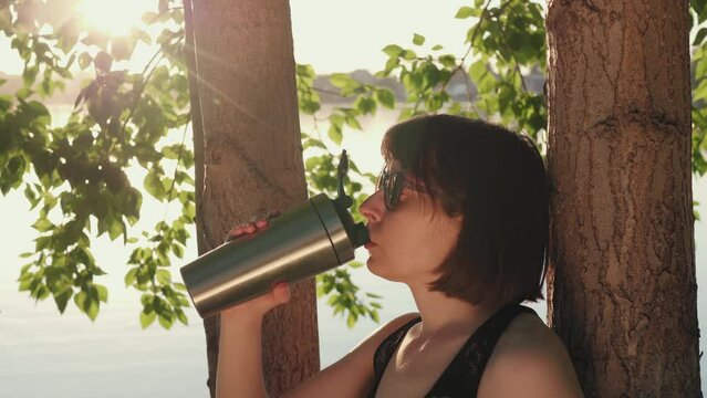 Sporty brunette woman in sunglasses drinks from metal reusable bottle resting after workout under tree on the riverbank.