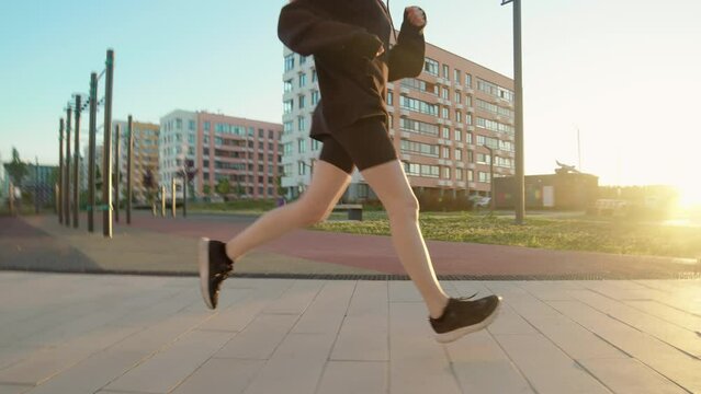 Handheld unrecognizable girl in black leggings and sweater is doing morning run along street with sports ground in background. Young woman running forward doing sports and workout outdoors
