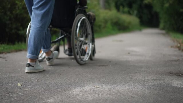 Unrecognizable Caucasian woman pushing wheelchair with African American man in summer park. Young girlfriend taking care of military boyfriend outdoors. Support and disability concept