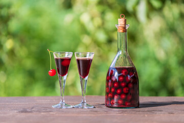 Homemade cherry brandy in two glasses and in a glass bottle on a wooden table in a summer garden