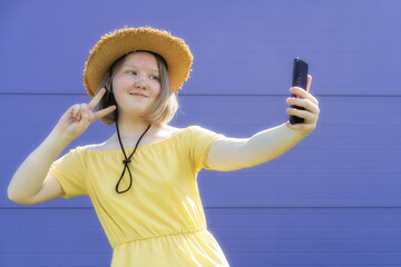 Asian teenager girl in yellow dress and hat shows greeting gesture while video chatting with friends on purple background. Trendy teenage culture. Generation Z