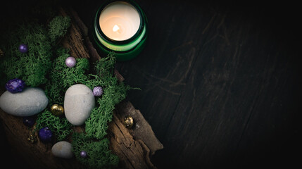 Fototapeta na wymiar Lilac, golden stone lies in the bark of a tree with green moss, gray stones and a burning candle on a black