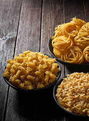 Three deep bowls with uncooked pasta on a wooden table. Assortment of raw macaroni
