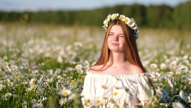 A beautiful redheaded girl is enjoying life in a field of daisies at sunset. A young woman with a wreath of daisies on her head smiles happily in nature.