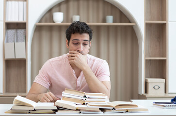 Upset young male student covers his face with hand and sits at the table with a lot of books....