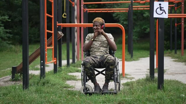 Wide shot portrait of positive brave soldier in wheelchair listening to music in headphones smiling. Courageous African American handsome young man enjoying leisure at sports ground in park