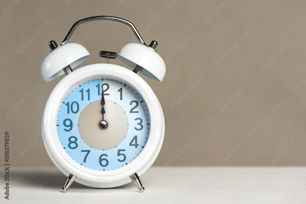 Poster twelve o'clock on the alarm. a white alarm clock is on a white table. the clock hand points to 12 o'