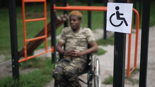 Disability accessibility sign on sports equipment in park with African American young man in wheelchair at background. Blurred confident soldier training outdoors on summer day. Lifestyle and sport
