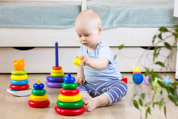 Cute baby boy toddler sitting on the floor at home and collects a pyramid toy.