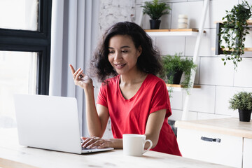Attractive young african american female sitting at table with cup, smiling looking at laptop screen, reading mail, chatting with friends, sharing social media, talking on video call